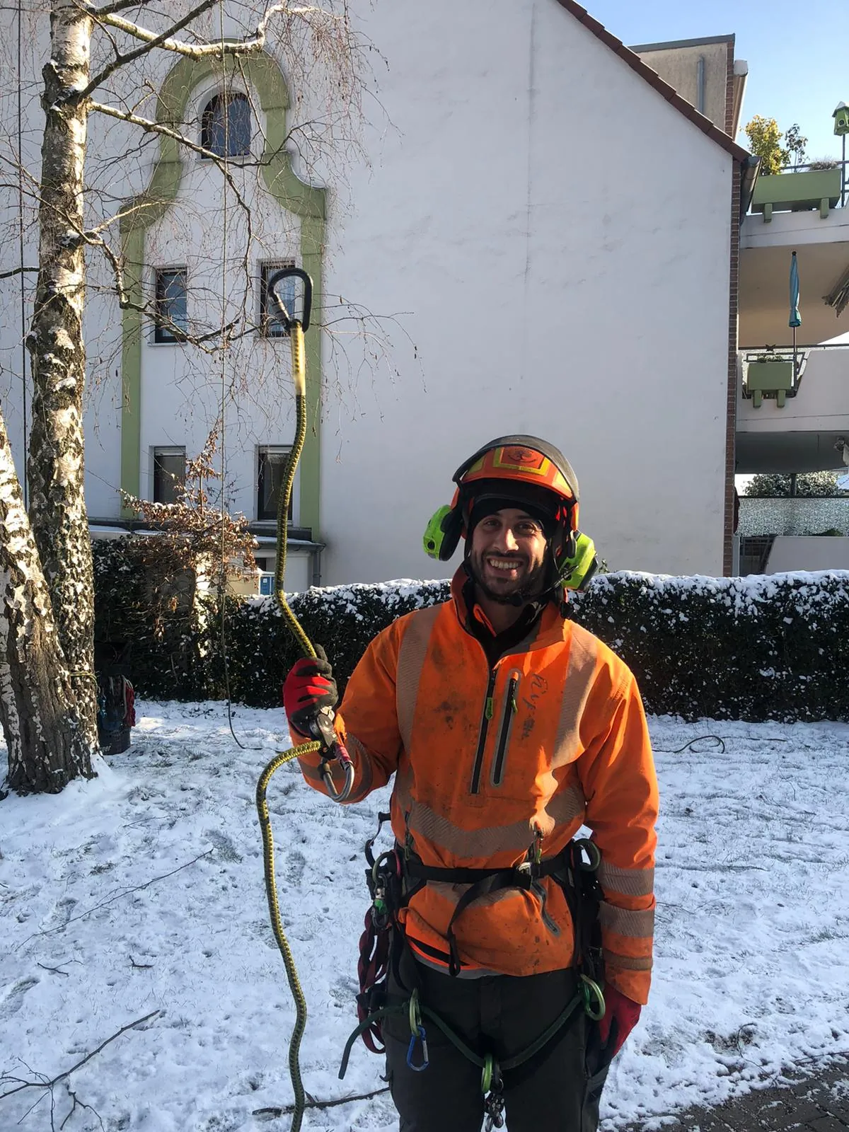 Arborista-Baumpfleger mit Kletterhelm, Gehörschutz und Klettergurt hält im Schnee ein Kletterseil mit Karabiner
